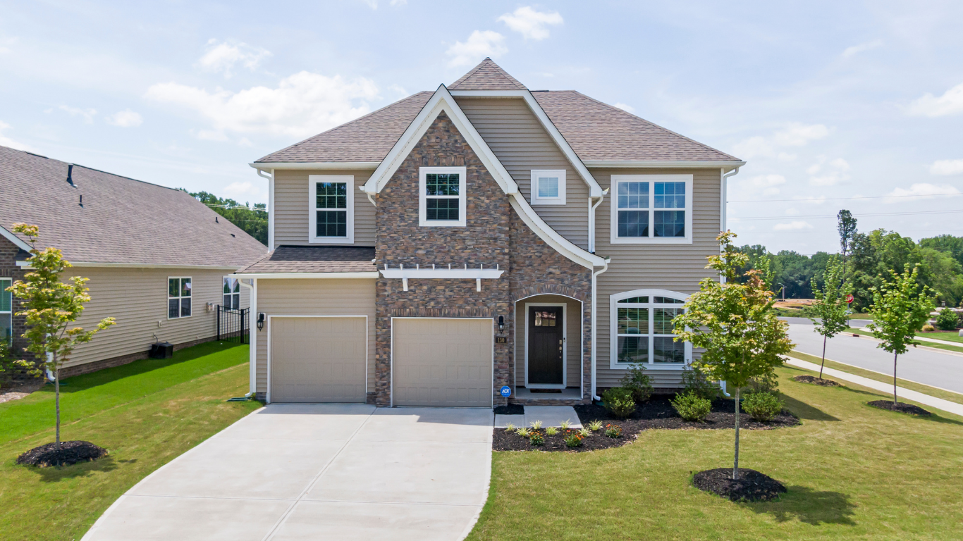 Two-story beige house with stone accents, a driveway, and a garage. It is daytime with blue skies and a lawn.