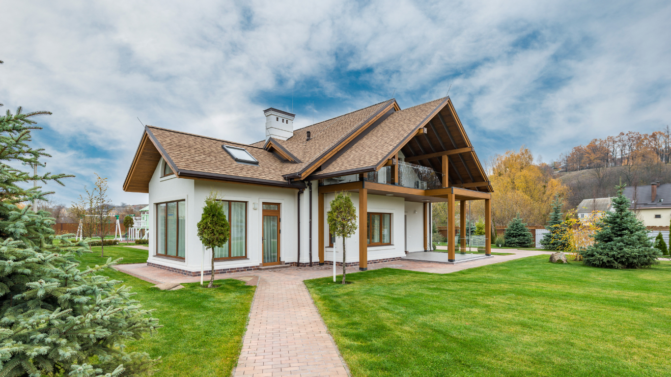 White house with brown roof and trim, surrounded by green lawn and trees under a cloudy sky.