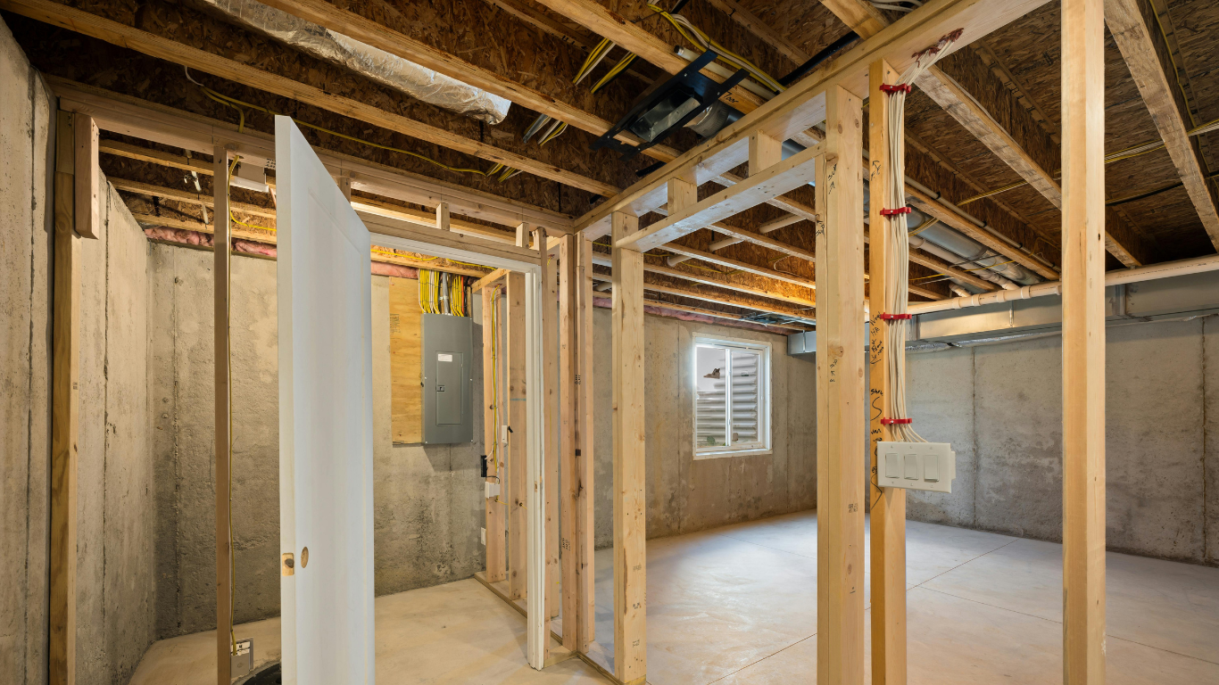 Basement interior under construction: bare wooden framing, concrete walls, unfinished ceiling, electrical panel.