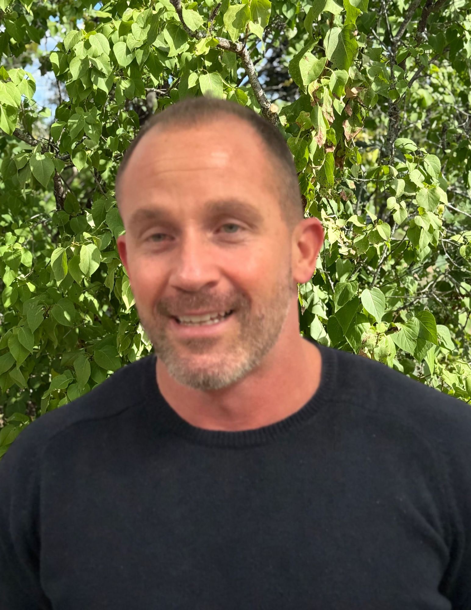 A man with a beard wearing a black sweater is smiling in front of a white brick wall.