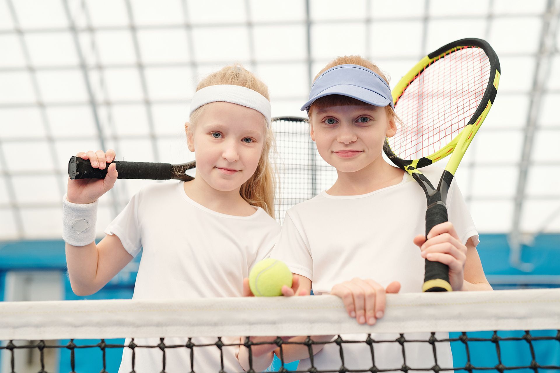 Two young girls are holding tennis rackets and a tennis ball on a tennis court.