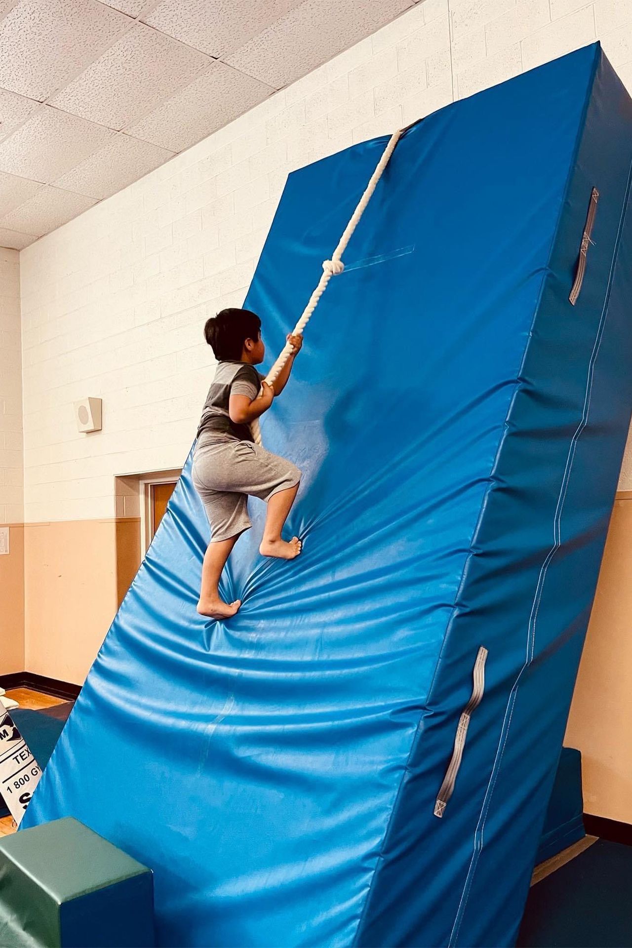 A young boy is climbing a blue mat with a rope.