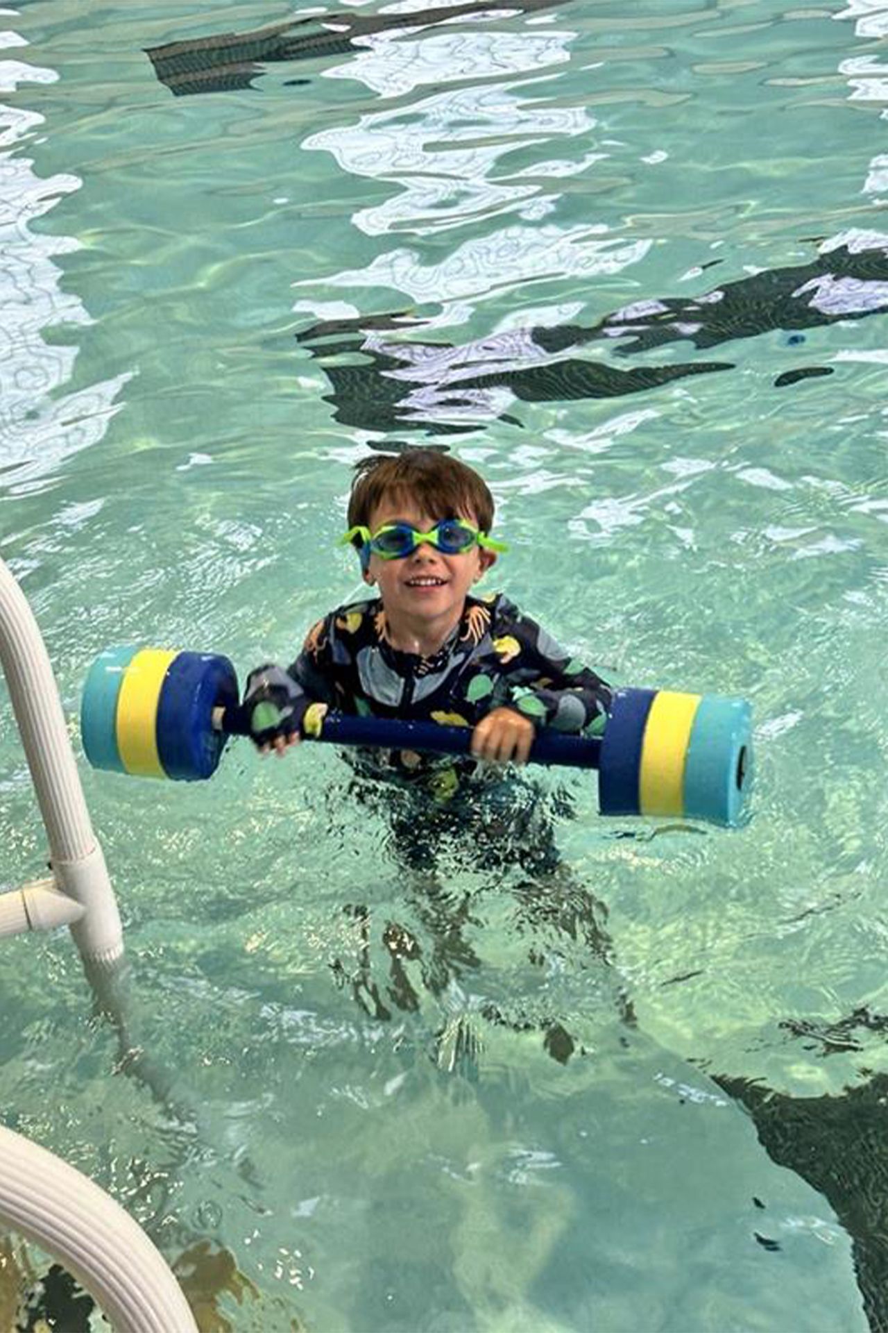 A young boy is swimming in a pool with a foam dumbbell.