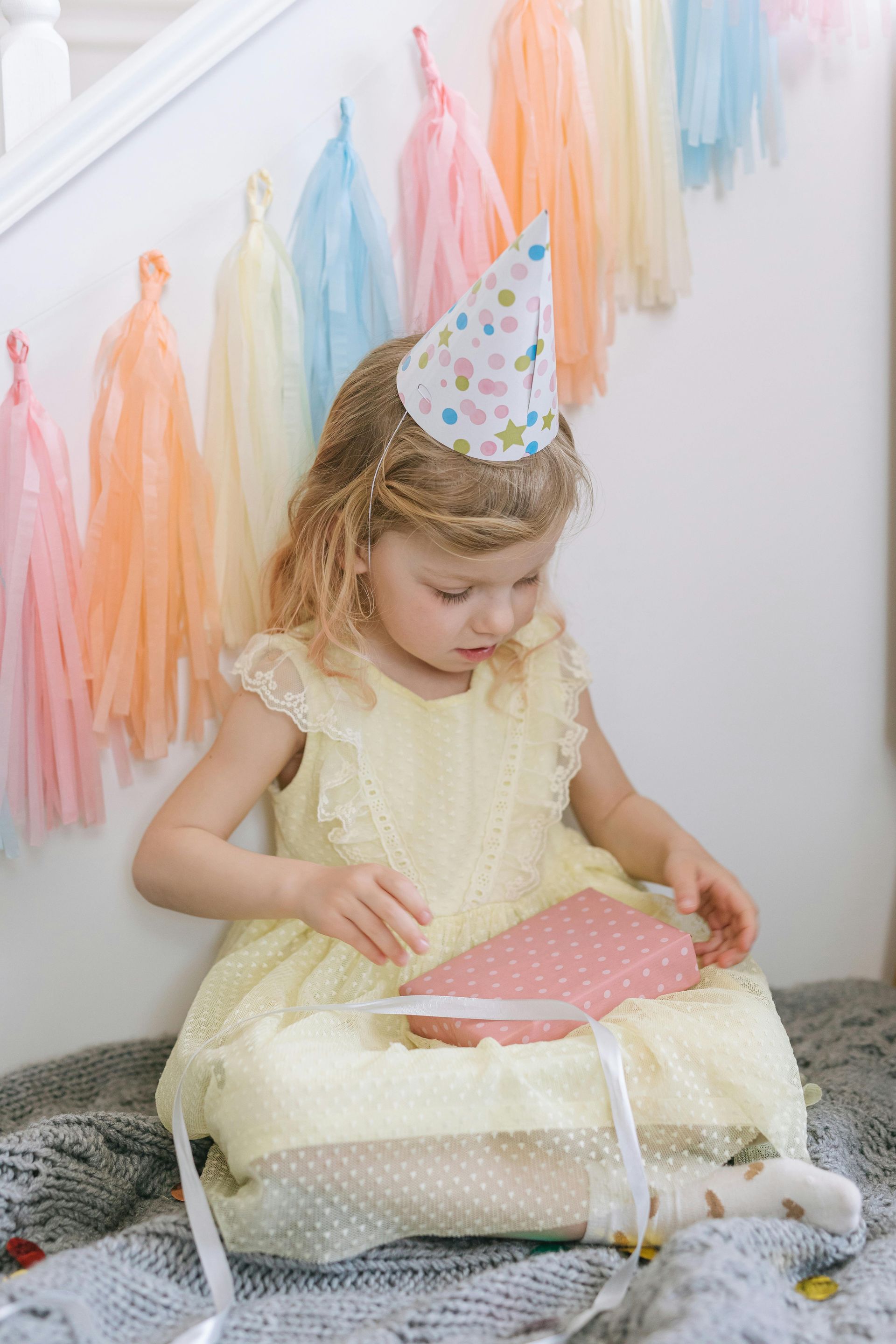 A little girl in a yellow dress and party hat is sitting on the floor.
