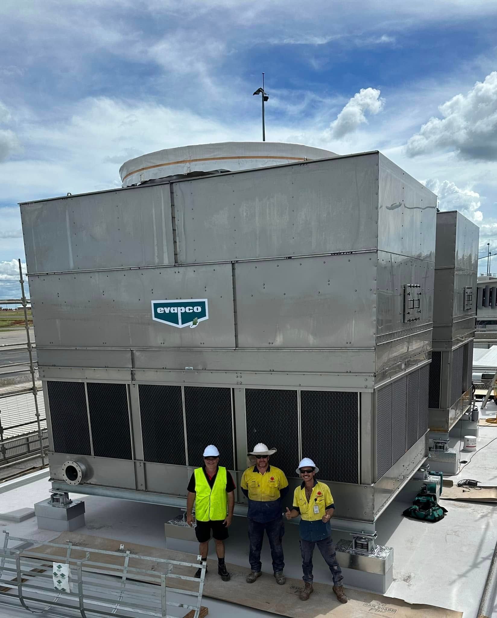 Three workers stand in front of a large industrial cooling tower on a rooftop, under a cloudy sky — Great Northern Airconditioning & Mechanical Services in Berrimah, NT
