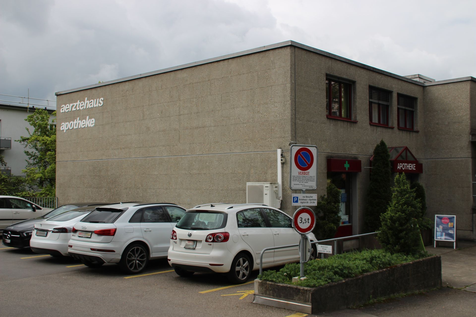 Gebäude mit geparkten Autos; Schild mit der Aufschrift „Apotheke“.