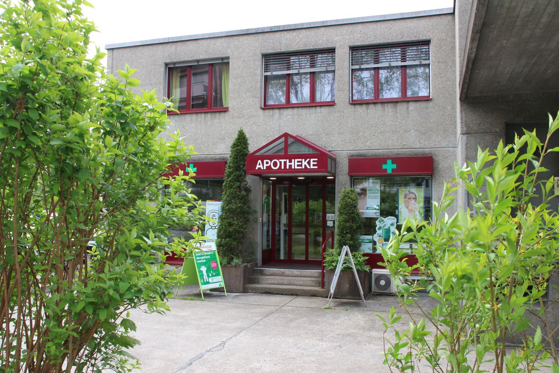 Aussenansicht einer Apotheke, Schild „APOTHEKE“ über der Tür, grünes Kreuzlogo, kleines Gebäude, Grün davor.