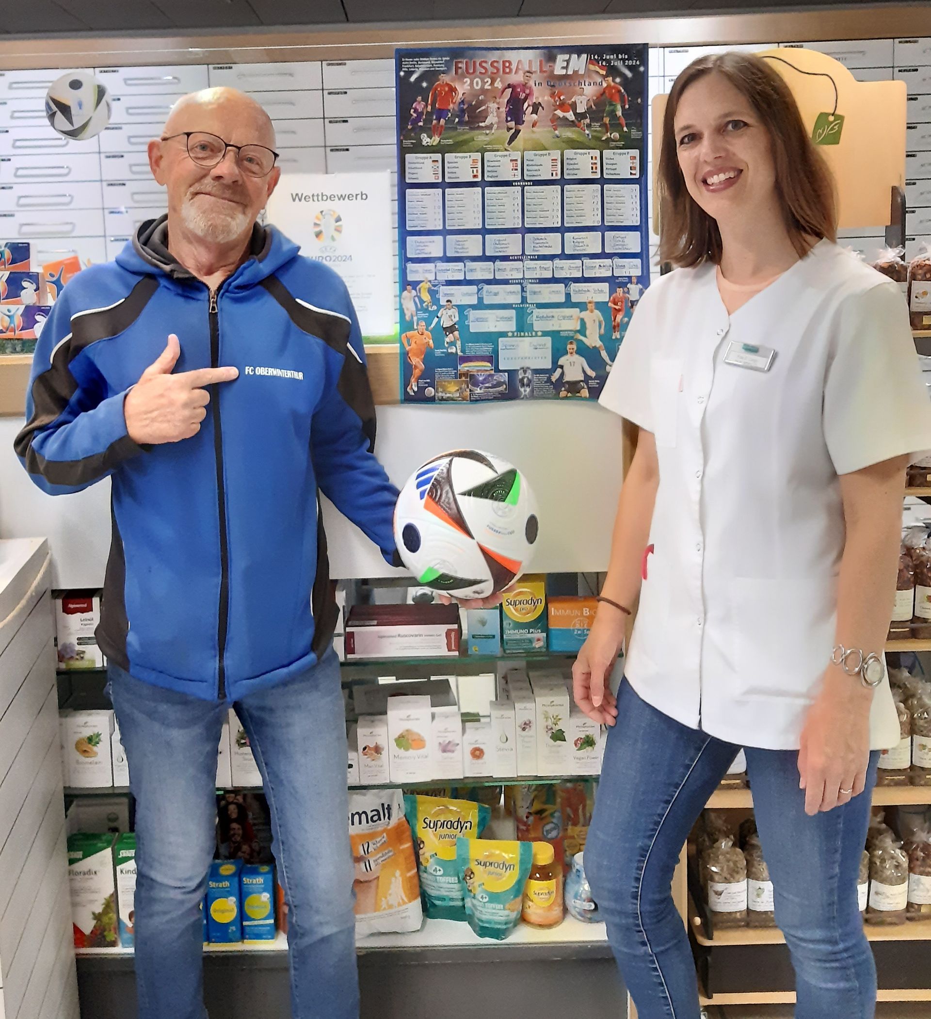 Ein Mann in blauer Jacke zeigt in einem Geschäft auf einen Fussball, den eine Frau in weissem Mantel hält.