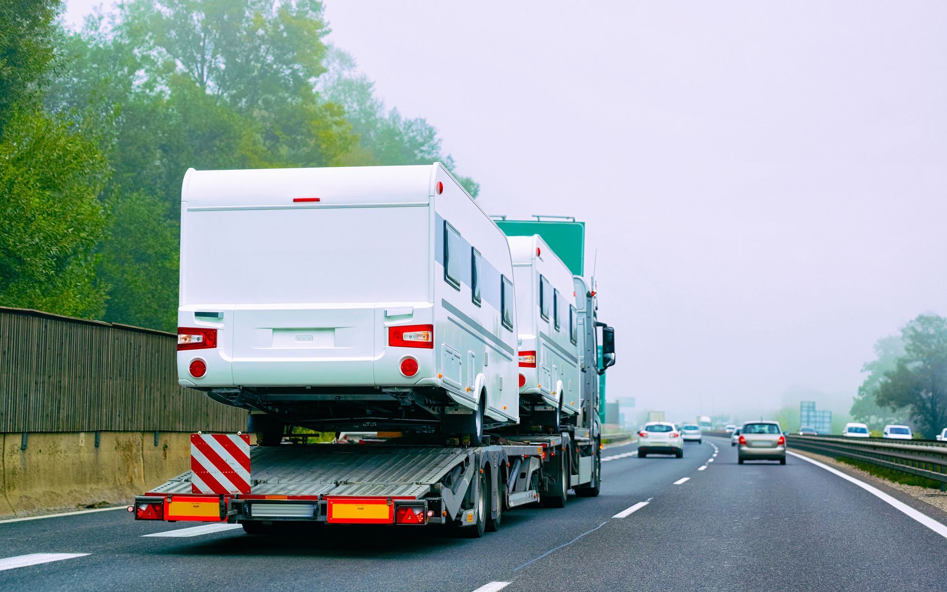 Semi-truck carrying two white RVs on a highway, overcast sky.