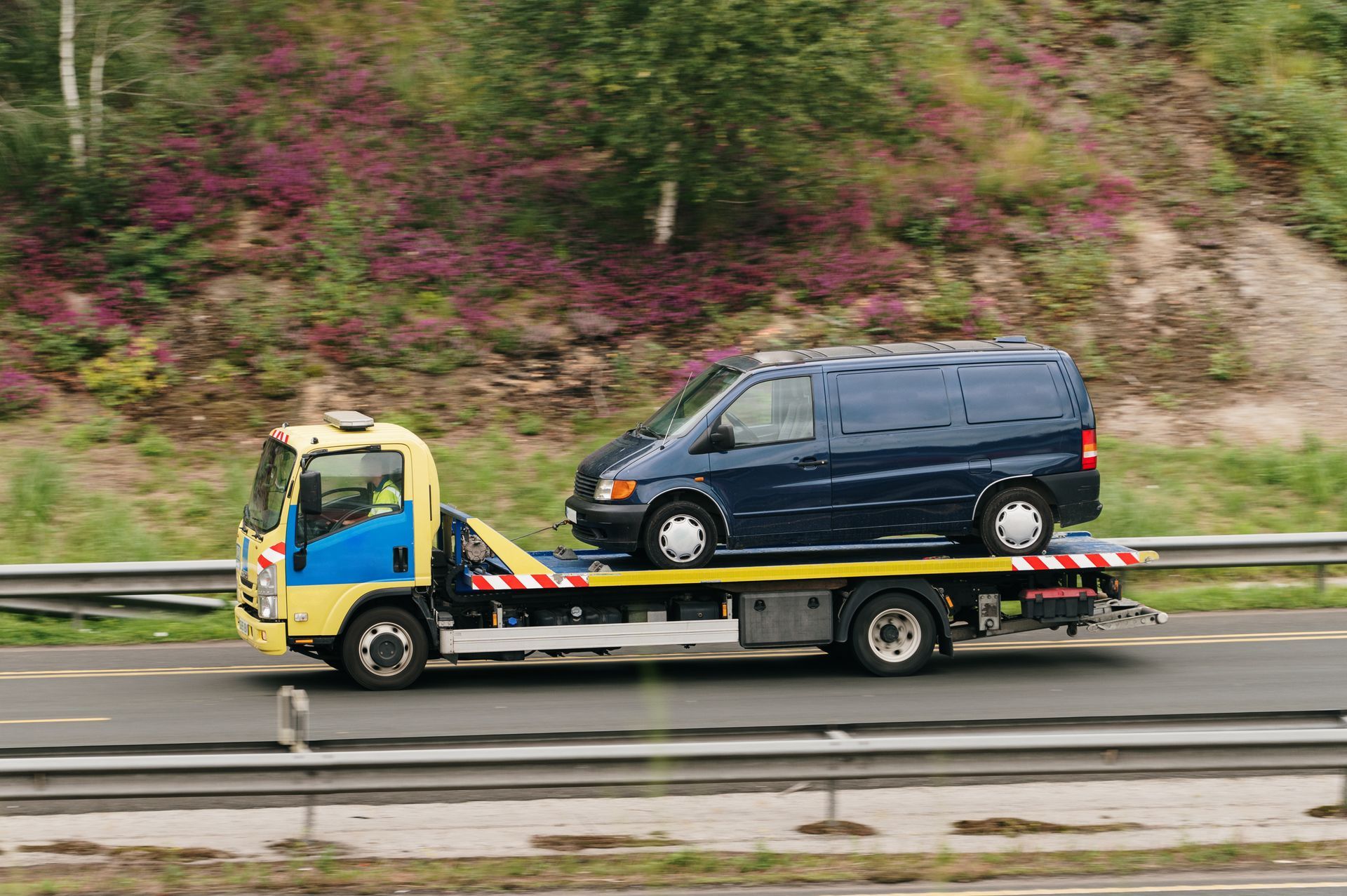 Yellow tow truck carrying a blue van on a highway, with blurred green and purple foliage in the background.