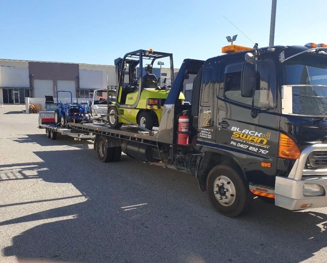 Black tow truck carrying a green forklift and blue equipment trailer on a sunny day.