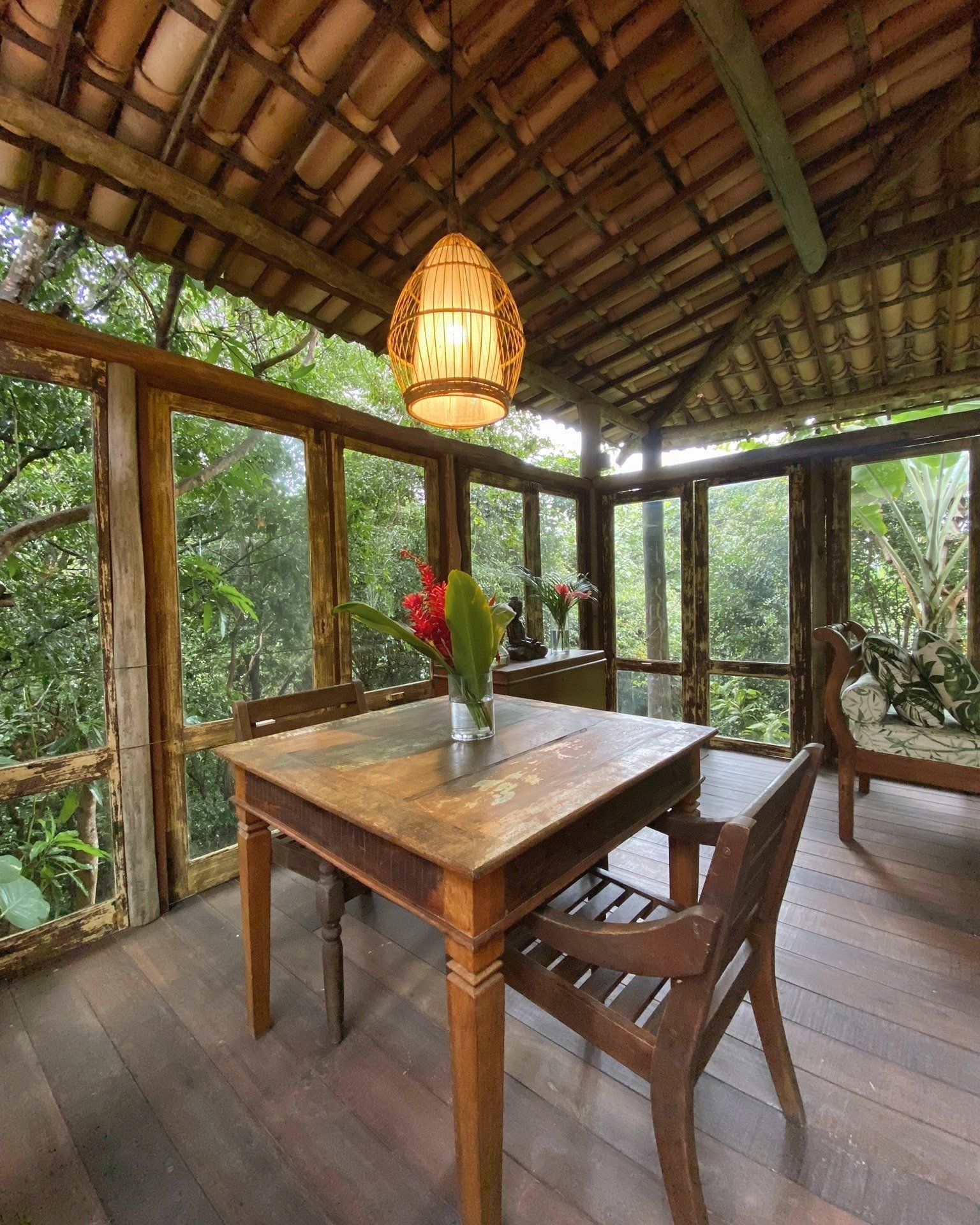 Uma mesa de madeira com um vaso de flores em cima dela em uma sala com muitas janelas.