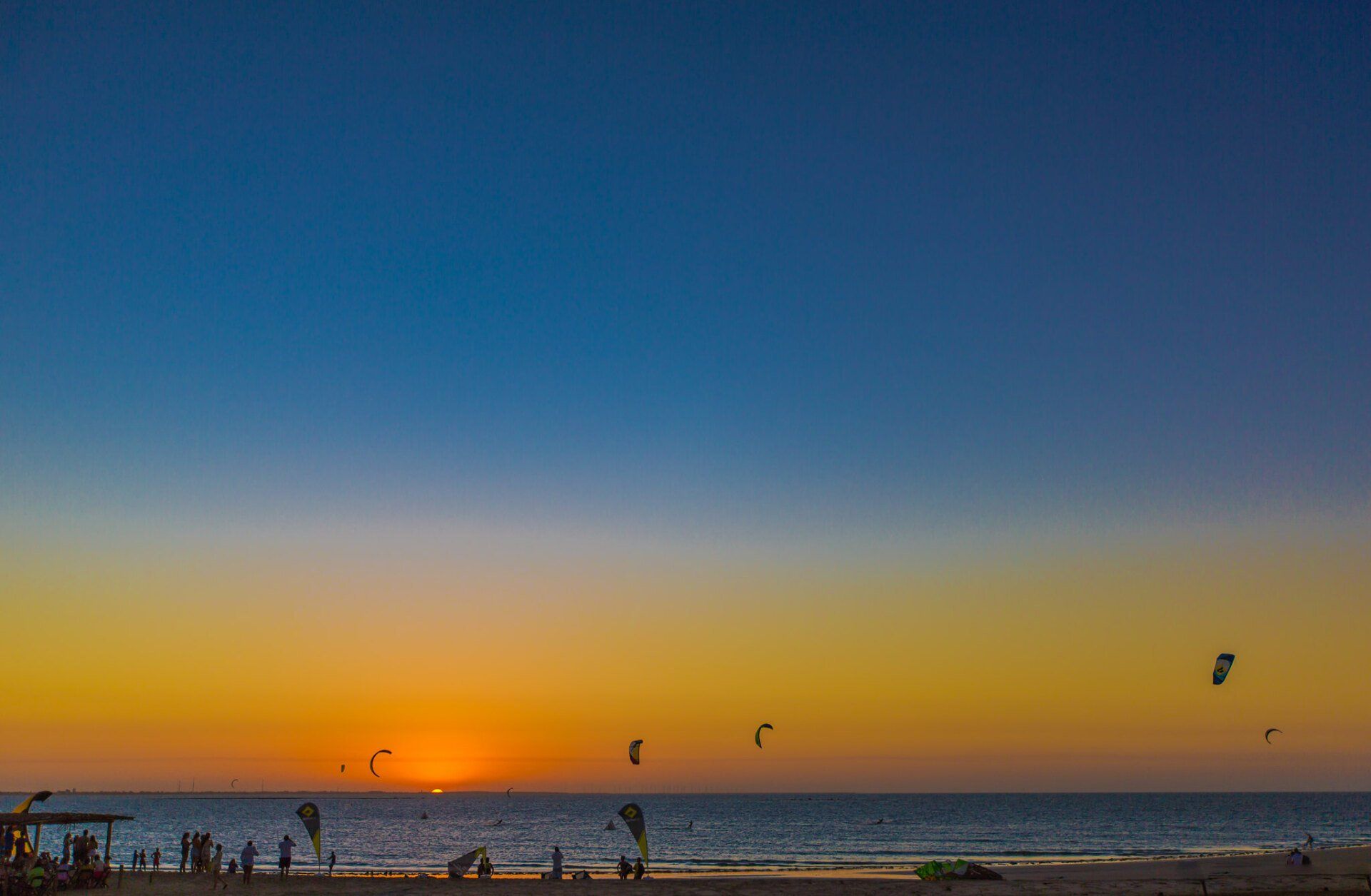 Um grupo de pessoas está empinando pipas em uma praia ao pôr do sol.