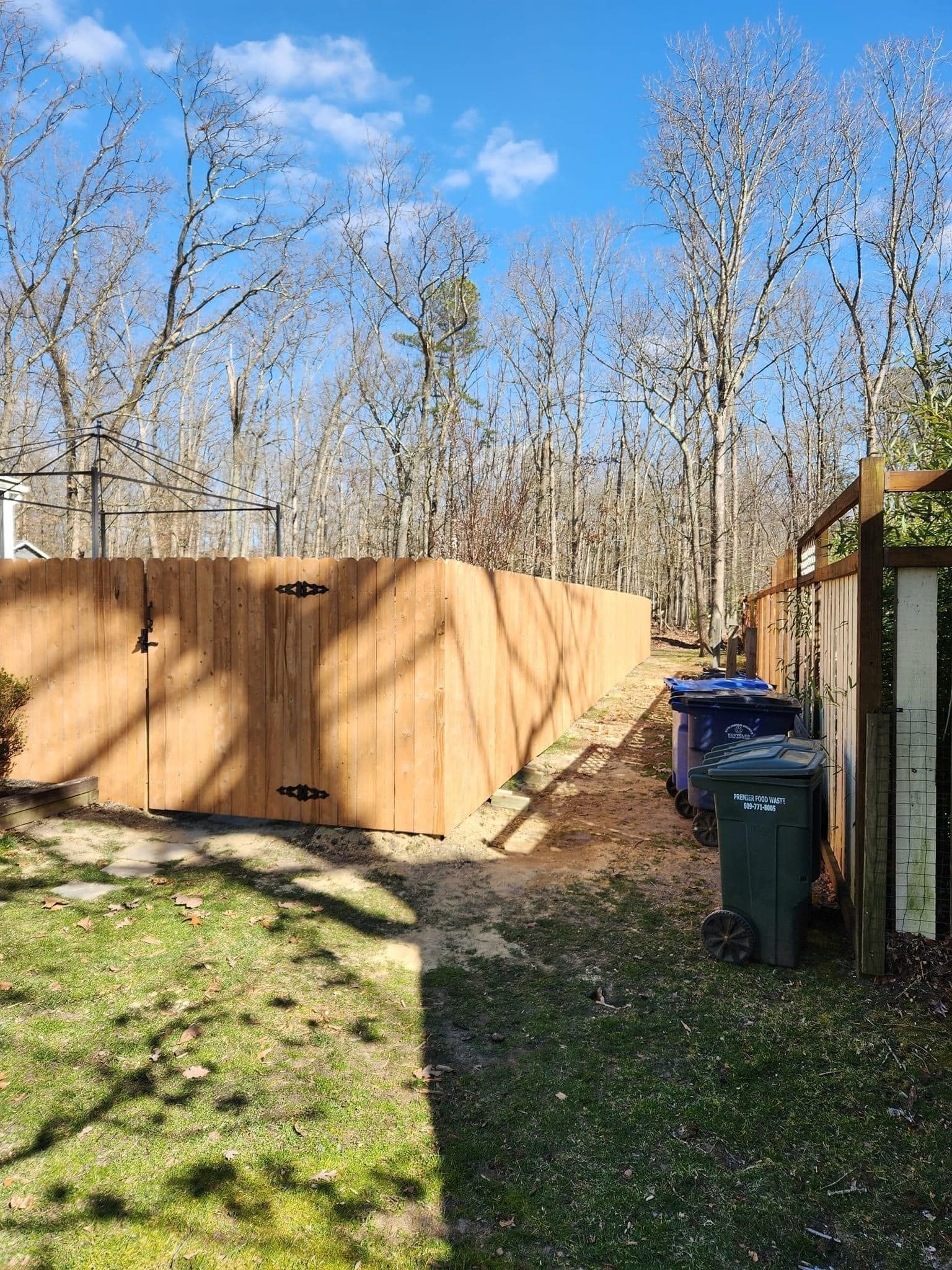 A backyard with a wooden fence, trash cans, and trees under a blue sky. A backyard with a wooden fence, trash cans, and trees under a blue sky.