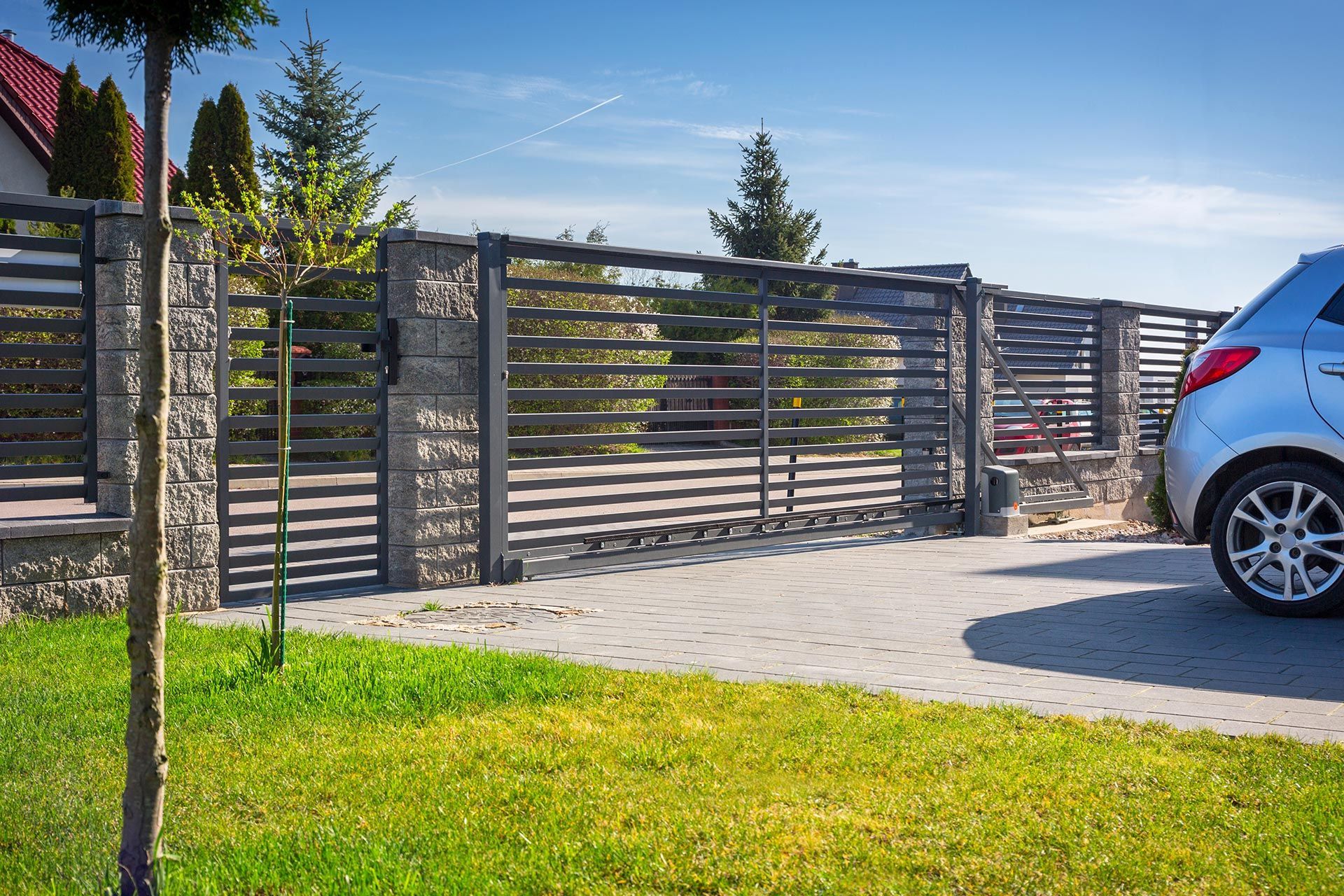 Gray car parked in front of a gray horizontal-slat gate and stone pillars on a paved driveway; green lawn and blue sky. Gray car parked in front of a gray horizontal-slat gate and stone pillars on a paved driveway; green lawn and blue sky.