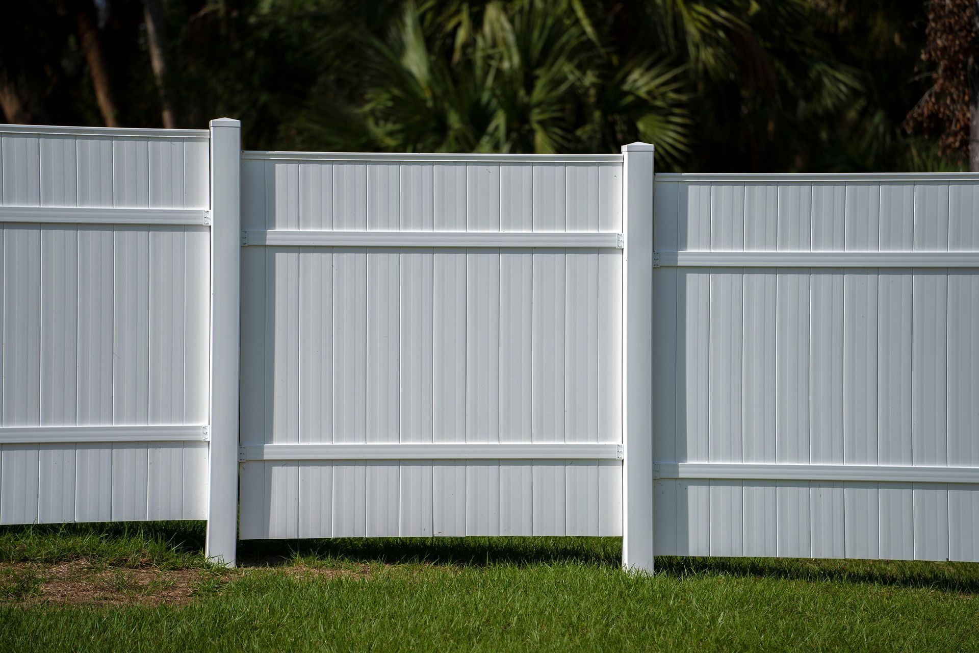 White vinyl privacy fence panels installed in segments with posts, positioned over a lawn with trees in the background.