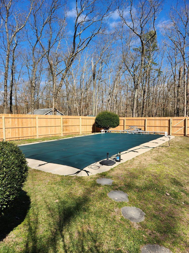 Pool covered with dark green tarp in a fenced backyard, surrounded by trees under a blue sky.