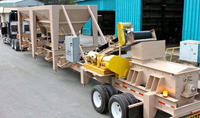 A trailer with a yellow machine on it is parked in front of a blue building.