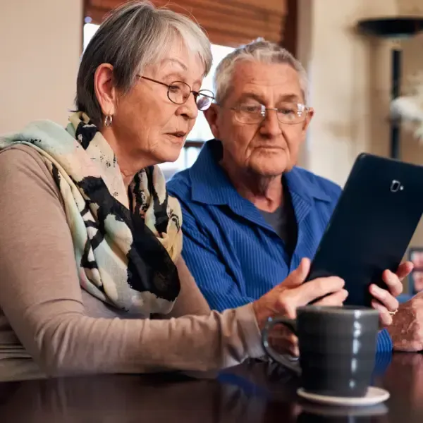 An elderly couple sitting together indoors, attentively looking at a tablet. The woman wears glasses and a scarf, while the man wears glasses and a blue shirt, holding the tablet. A coffee mug is visible in the foreground.