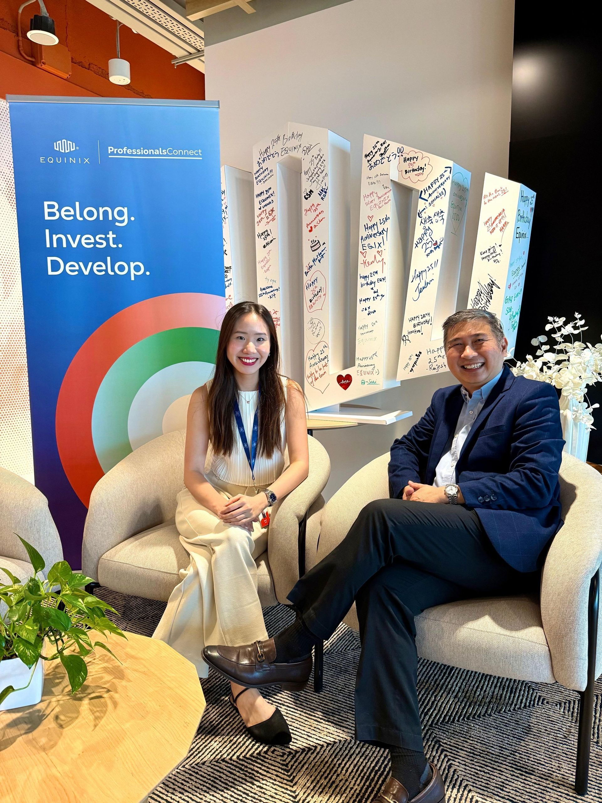 Two people seated, smiling, in front of a sign. Woman in cream outfit, man in navy blazer. Bright setting.
