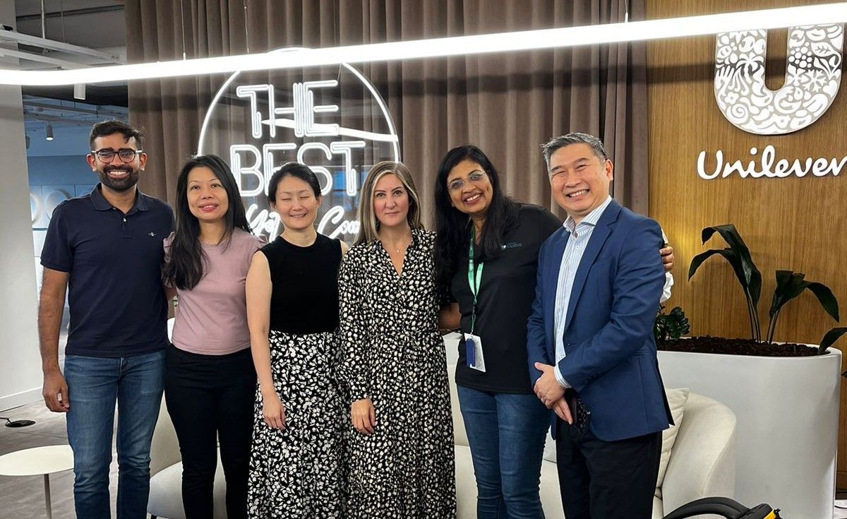 Six people posing for a photo in an office with Unilever branding. The group includes diverse people with smiles, likely colleagues.