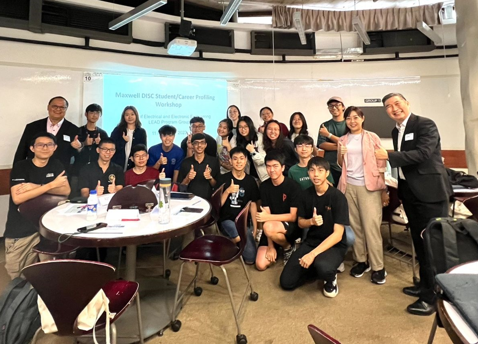 Group of students and instructors smiling for a photo in a classroom; a presentation is on the screen.