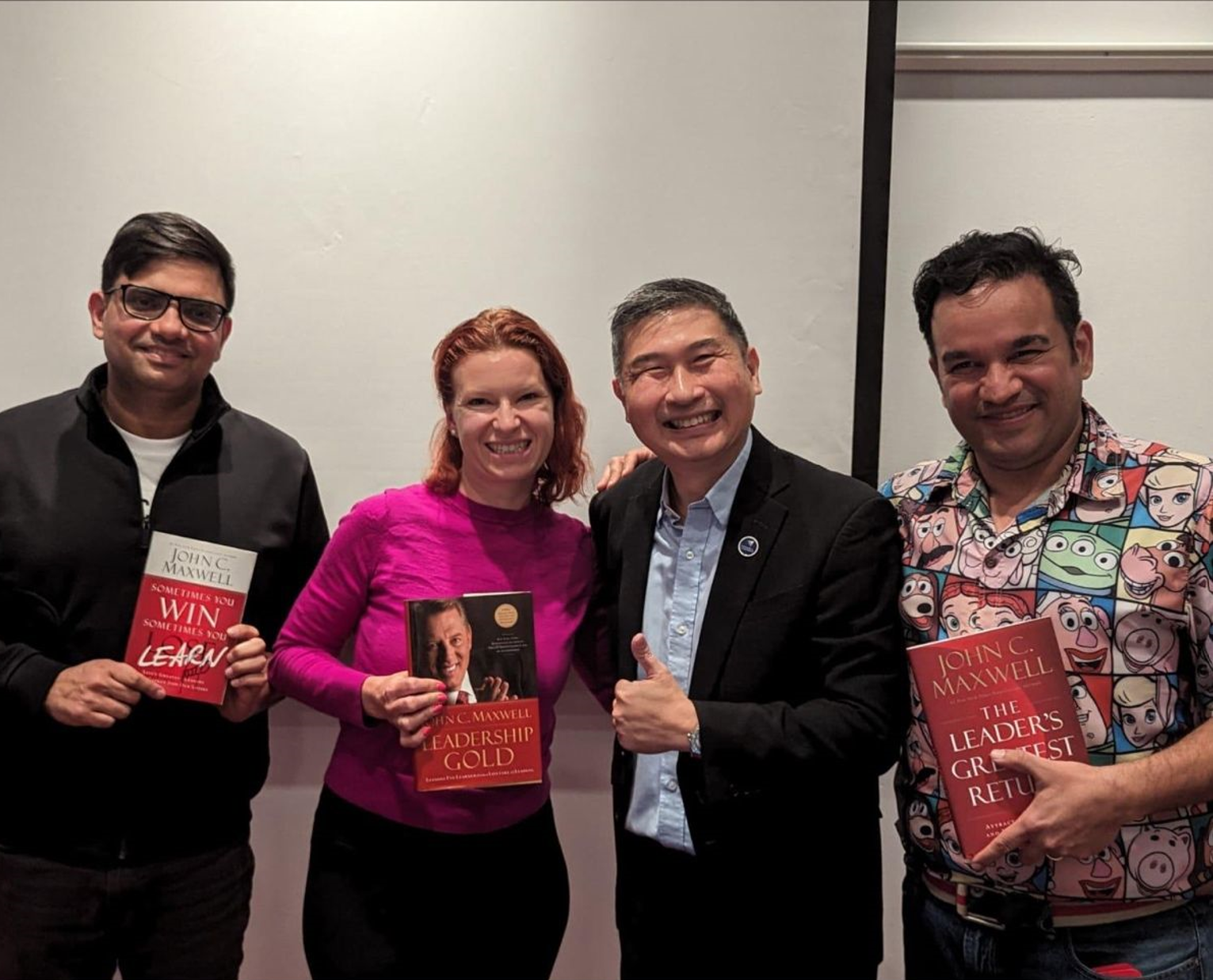 Four people pose with books; one man gives a thumbs-up. The setting is indoors with a white wall.