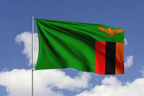 Zambian flag waving in the wind against a blue sky with clouds. Green field with vertical stripes and eagle.