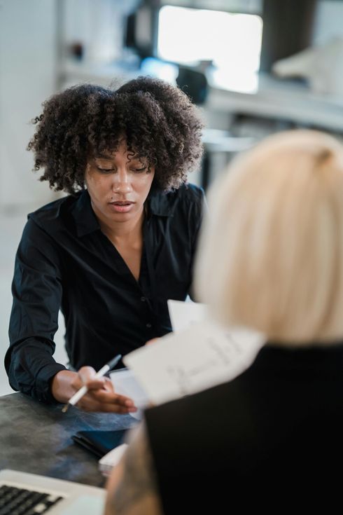 Woman with curly hair pointing at papers, speaking to another person in an office setting.