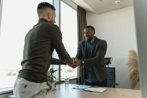 Two men shaking hands in an office, smiling. A desk, window, and papers are visible.