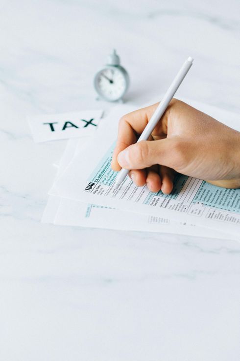 Person's hand filling out a tax form with a pen on a white surface next to a clock and 