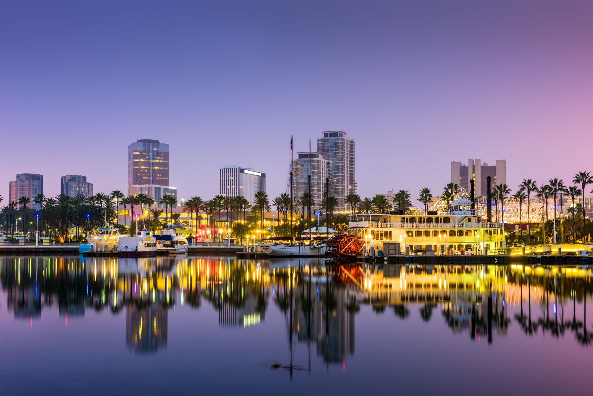 Long Beach, CA skyline at dusk reflected in calm water with palm trees.