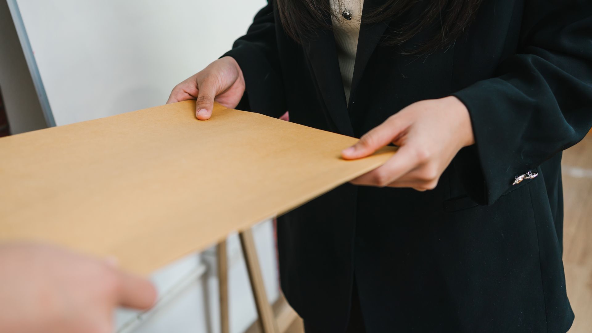 Person in a black blazer handing a brown envelope to another person.