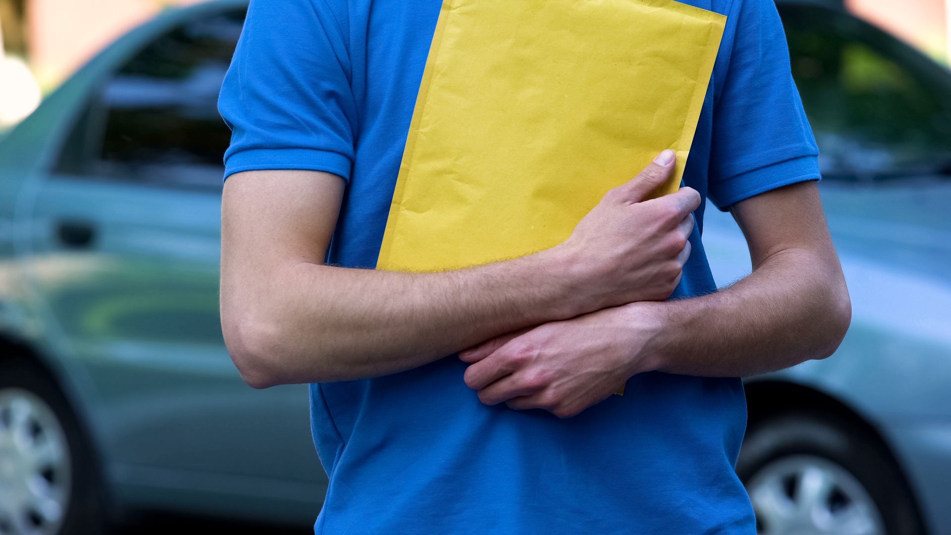 Person holding a yellow padded envelope, standing in front of a light blue car.