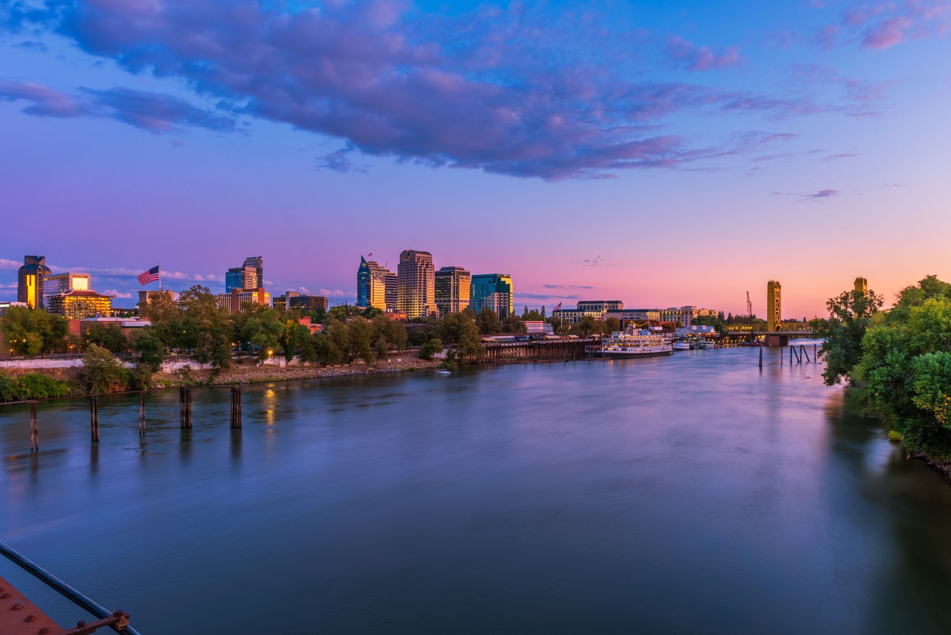 Riverfront cityscape at sunset; purple and pink sky reflects in the calm water.