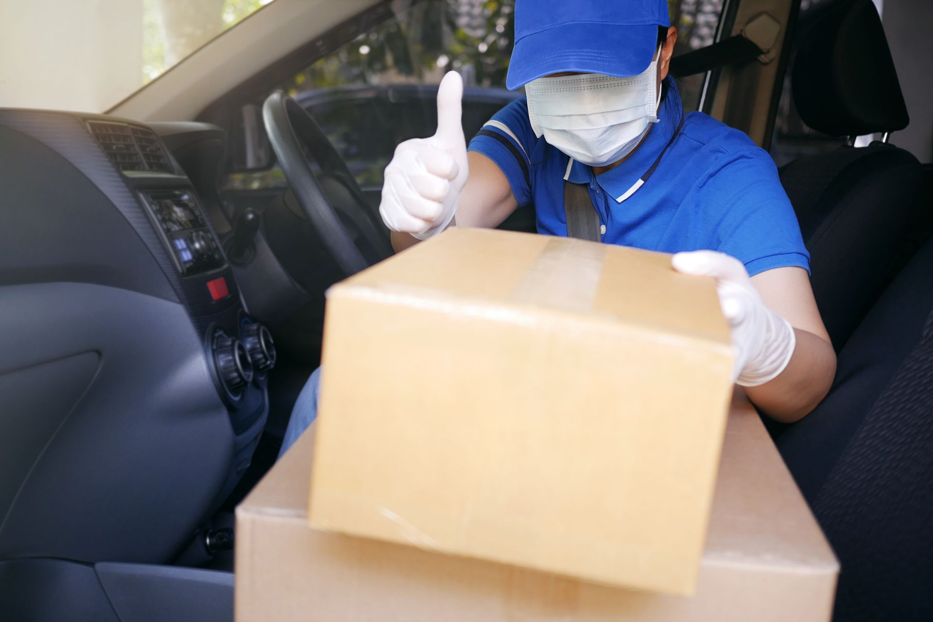 Delivery driver in car, wearing mask and gloves, giving a thumbs-up while holding a package.