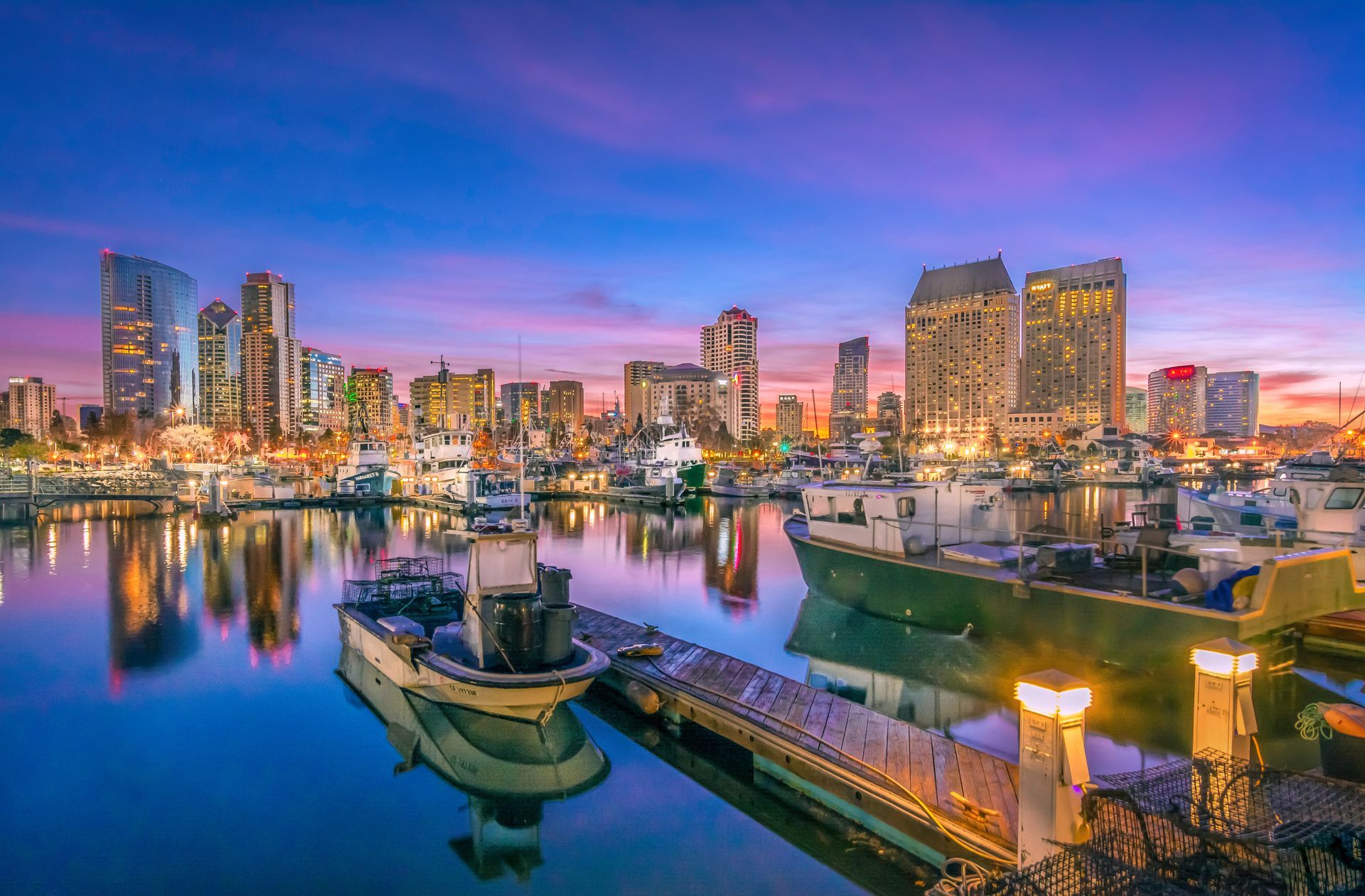San Diego skyline at dusk with boats in harbor; blue water reflects city lights and buildings against a purple and orange sky.