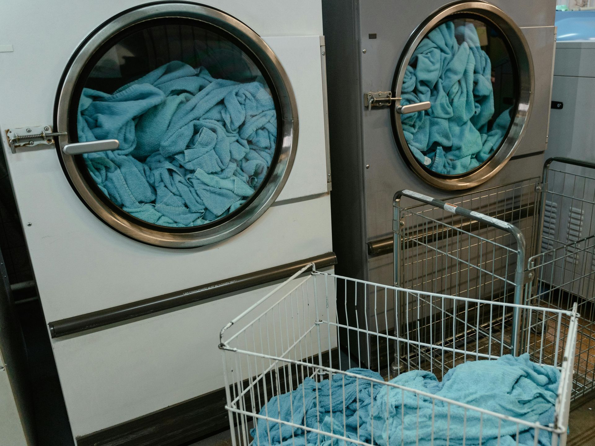 Two industrial clothes dryers with blue towels inside; a wire basket holds more blue towels.