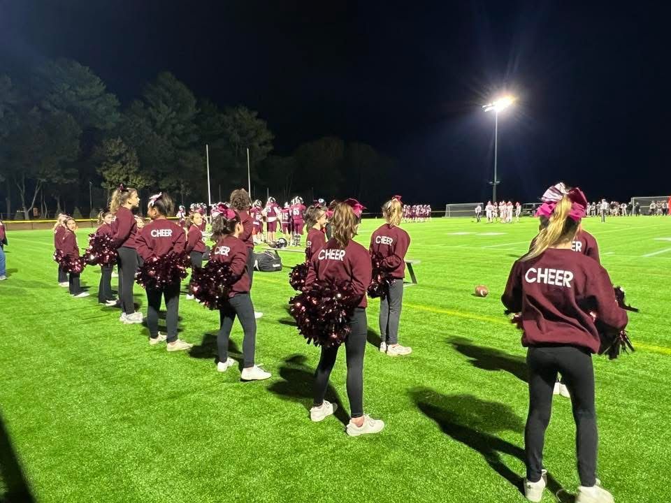 Cheerleaders in maroon uniforms practice on a lit football field at night