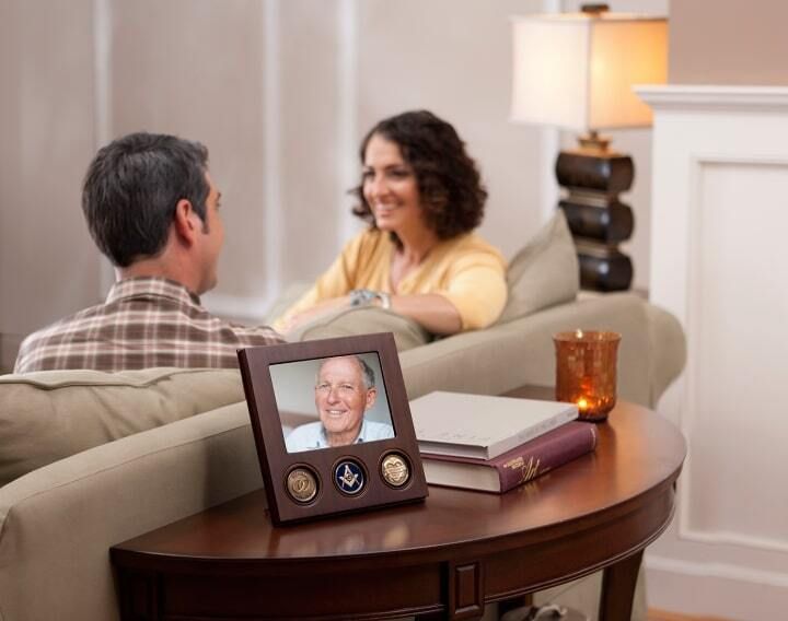 Couple on couch, smiling, with a framed photo of a man and military medals on a table.