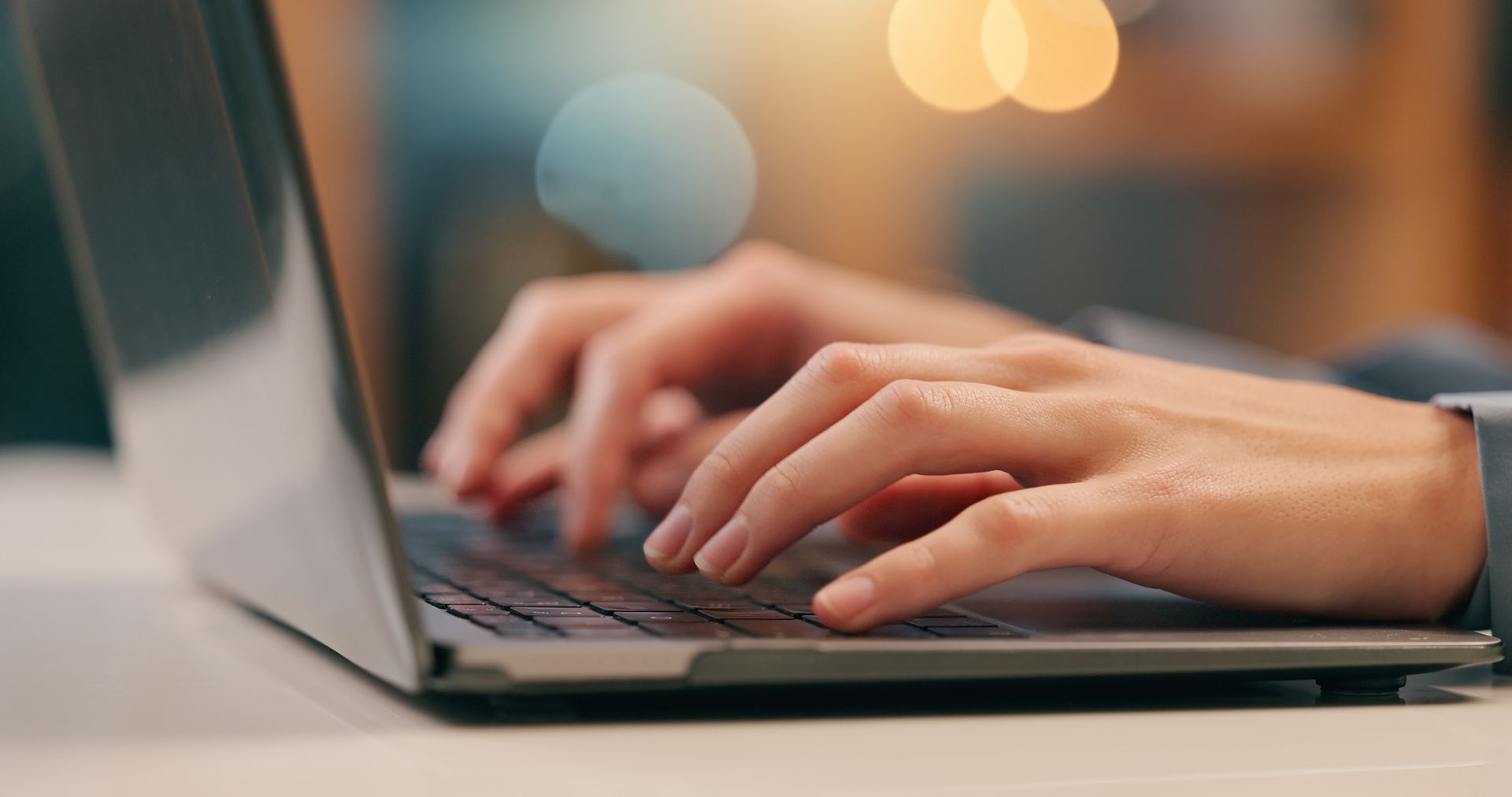Hands typing on a laptop keyboard; close-up. Blurry background with warm bokeh.