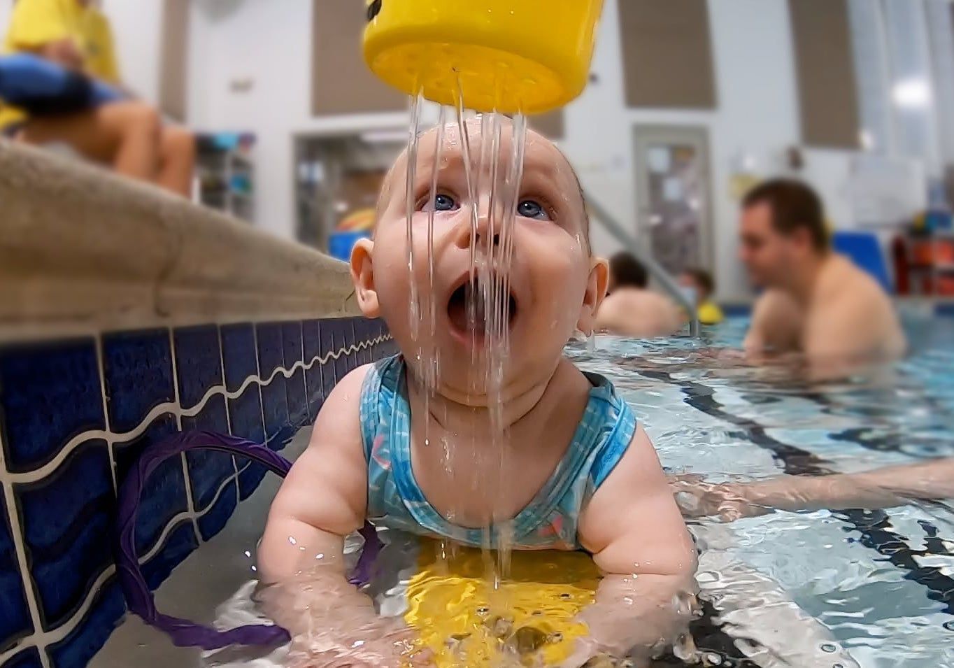 A baby is drinking water from a yellow bucket in a swimming pool.