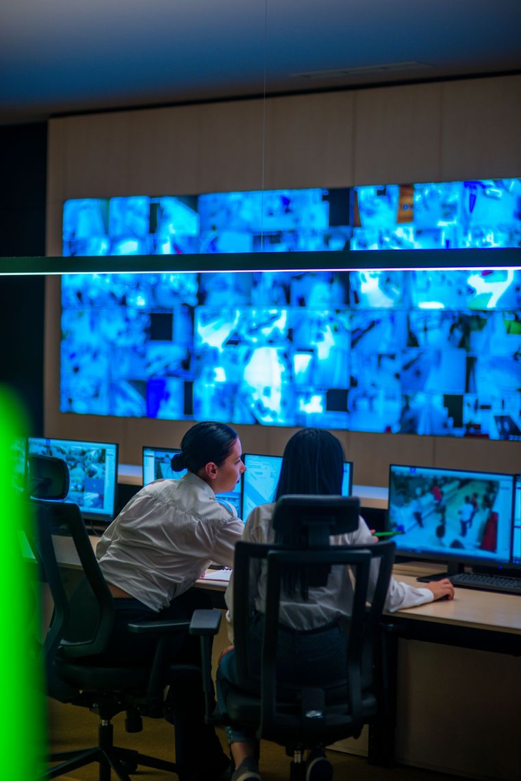 Two people monitoring screens in a security control room. Large wall display shows surveillance feeds.