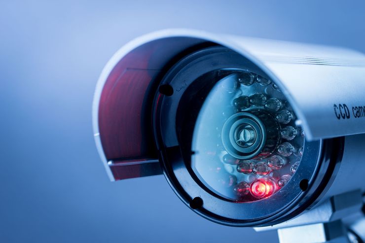 Close-up of a white security camera with a red light, against a blue background.