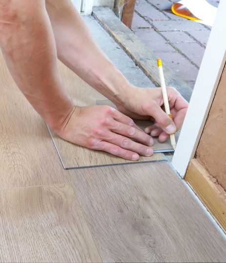 Hands measuring and marking a floor edge with a pencil beside a white door frame.