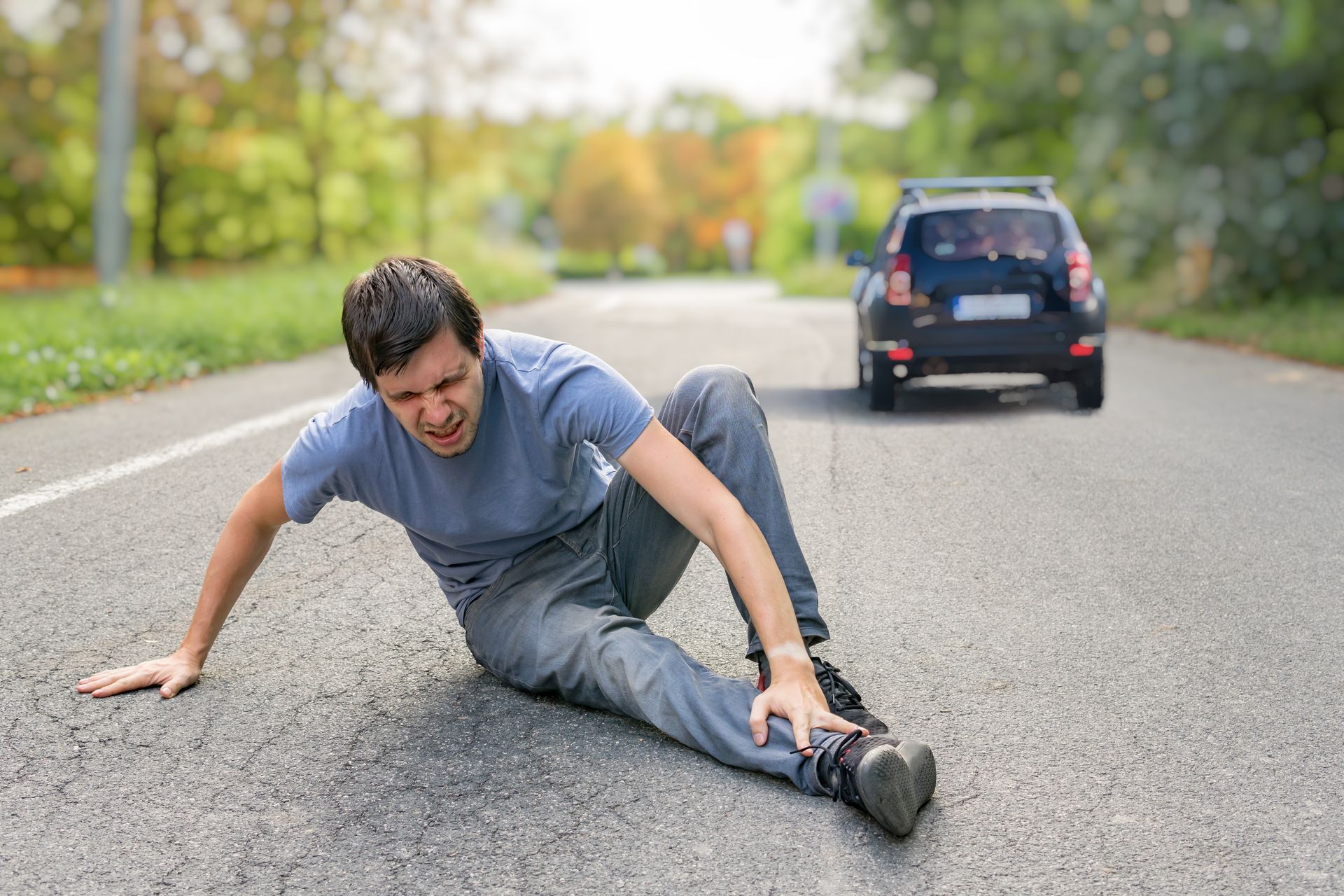 Man injured on road, grimacing, with car approaching in the background.