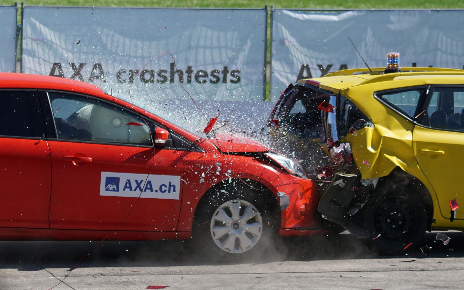 Two cars, red and yellow, collide during a crash test, water spraying, AXA crash tests in background.