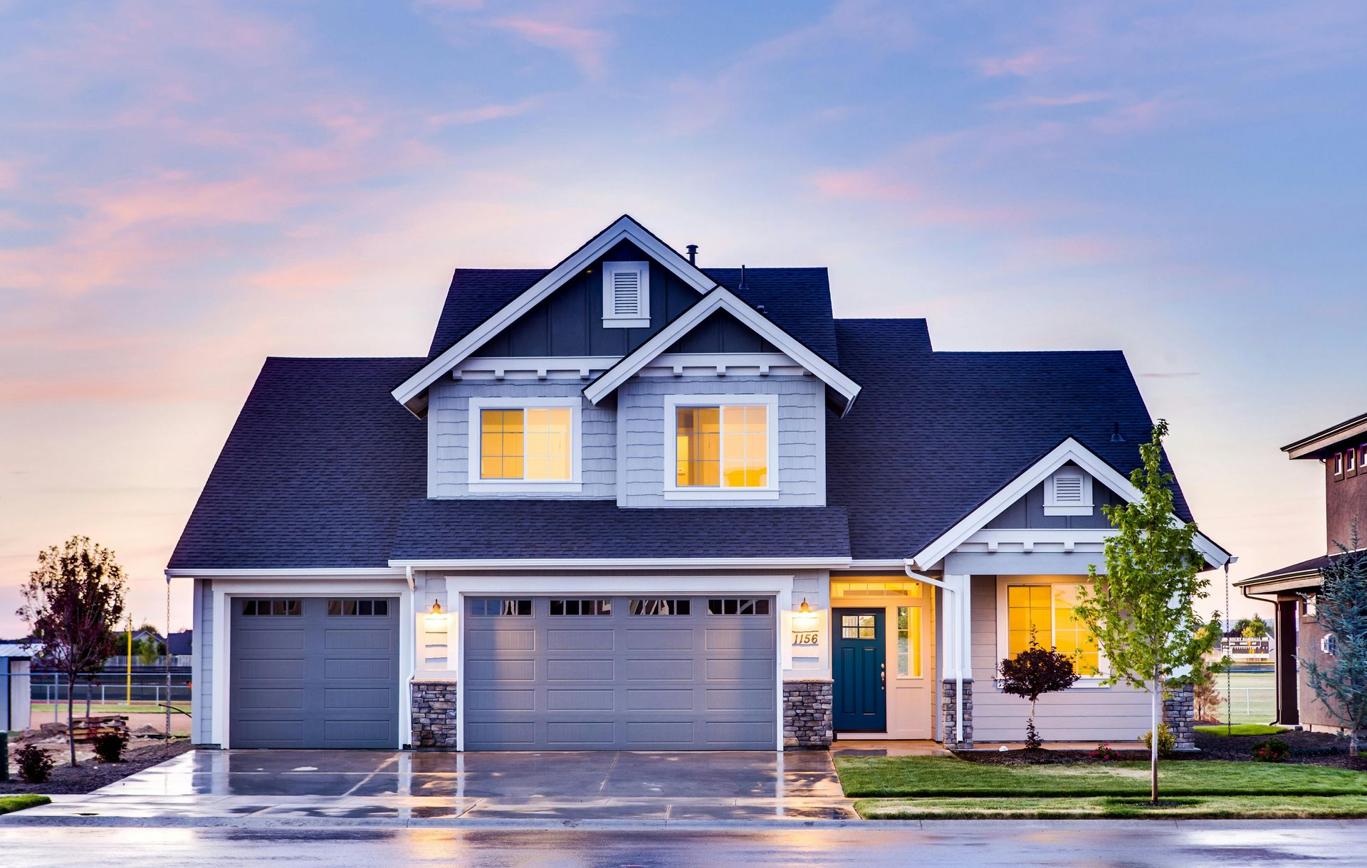 Two-story house with blue-gray siding, two-car garage, and a teal front door under a sunset sky.