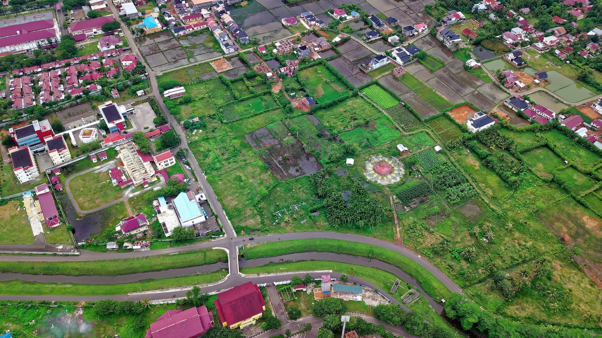 Aerial view of town with houses, fields, roads, and a bridge over a waterway. Green and brown predominate.