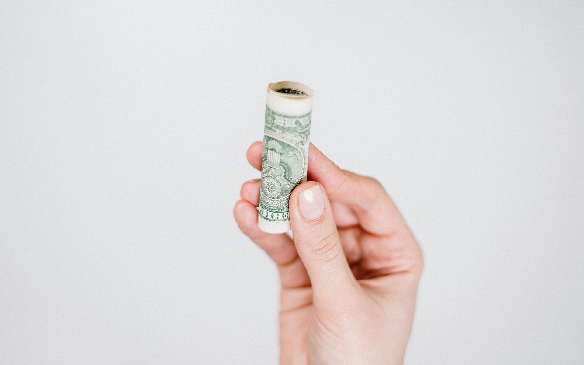 Hand holding a tightly rolled-up dollar bill against a plain white background.
